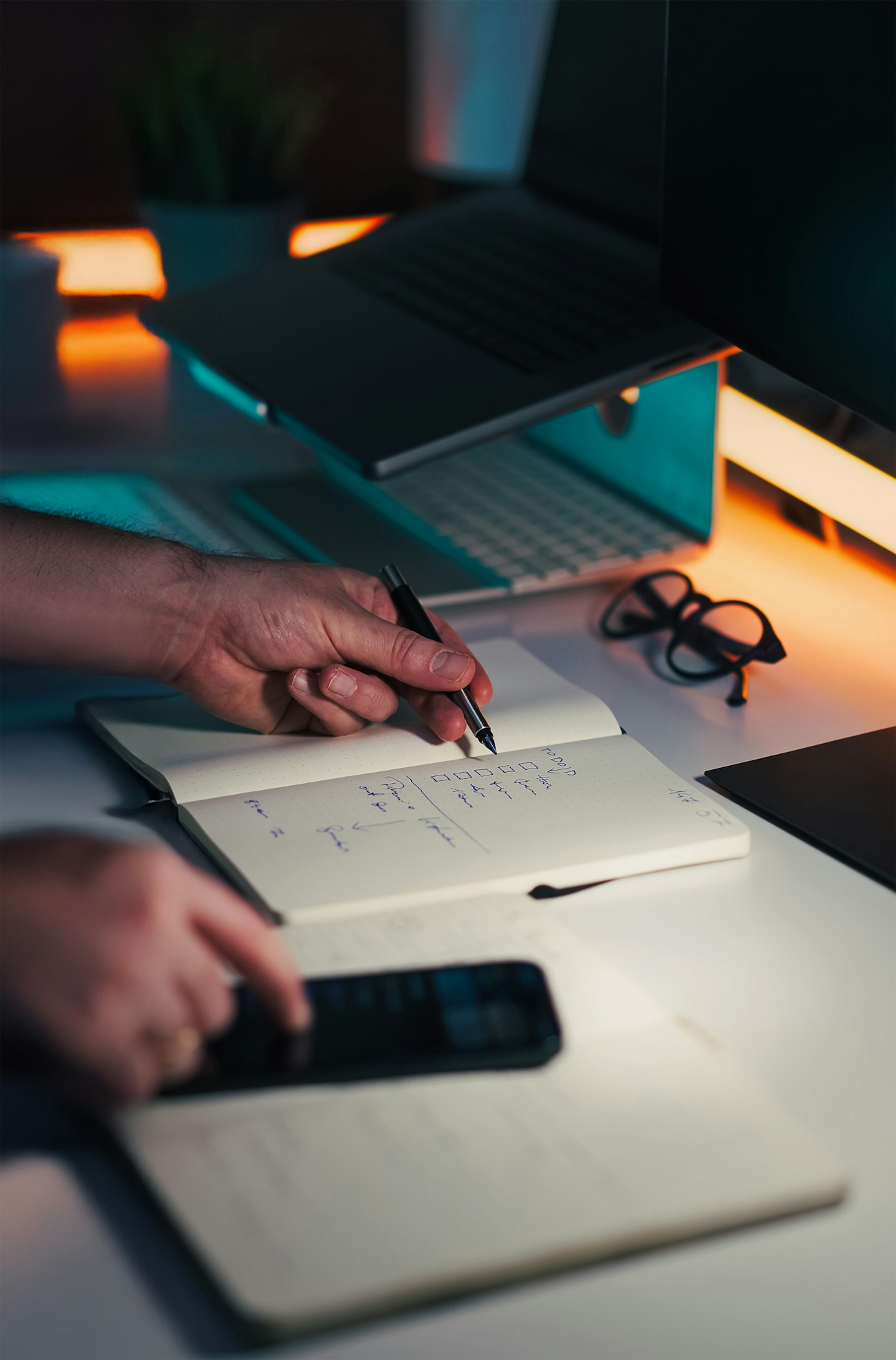 Mãos anotando ideias em um caderno junto a um notebook e smartphone